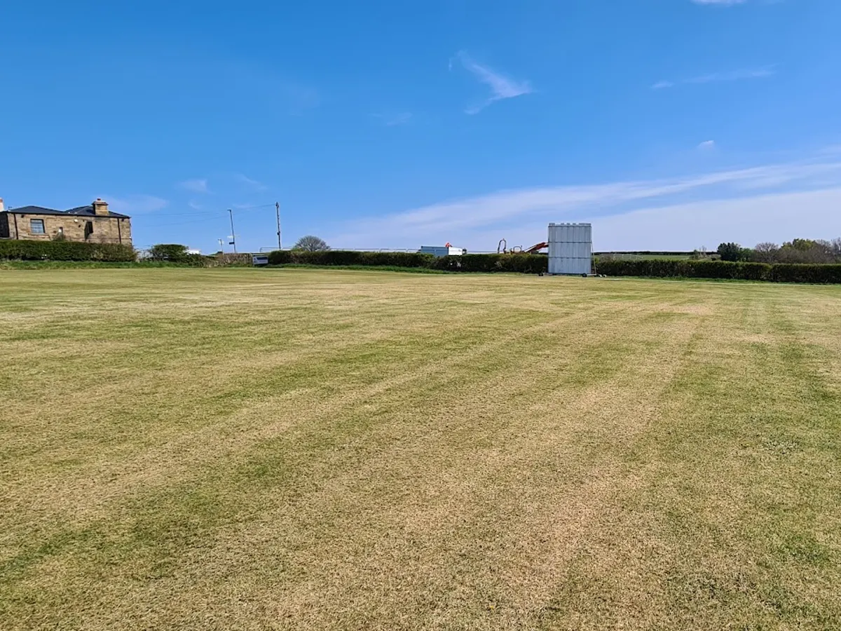 The cricket pavilion building with surrounding grounds, white and grey structure against a clear sky.