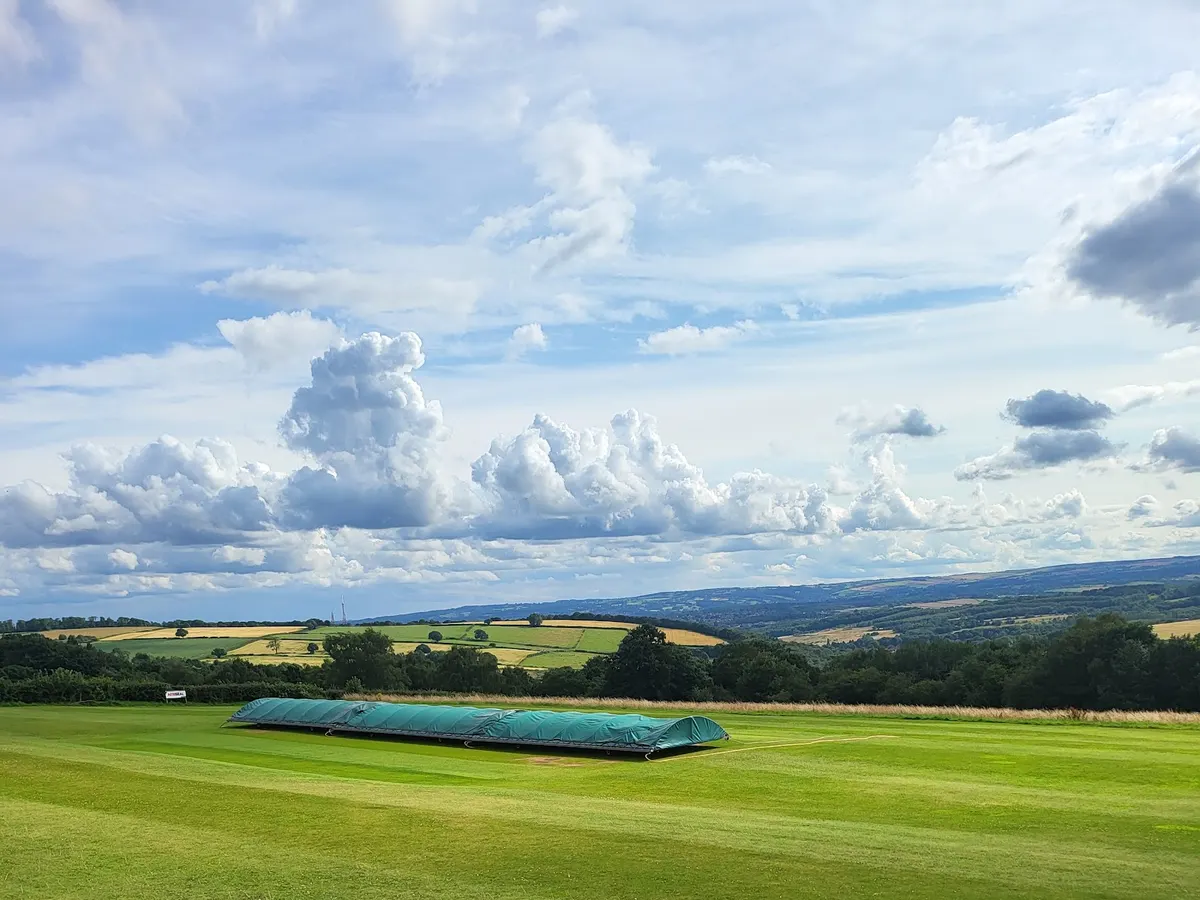 A covered cricket pitch with blue protective covers on the grass under a dramatic cloudy sky.