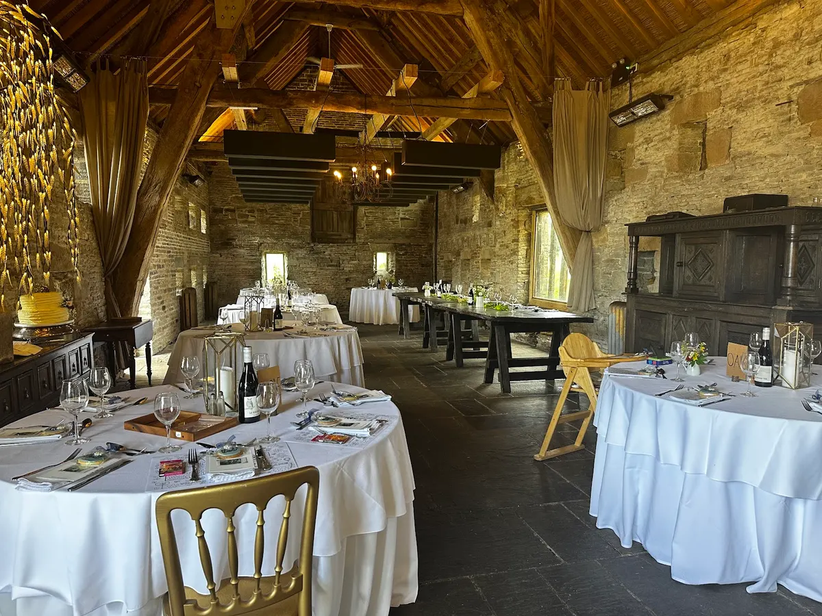 An elegant banquet hall with exposed wooden beams, round tables with white linens, brick walls, and candlelight.