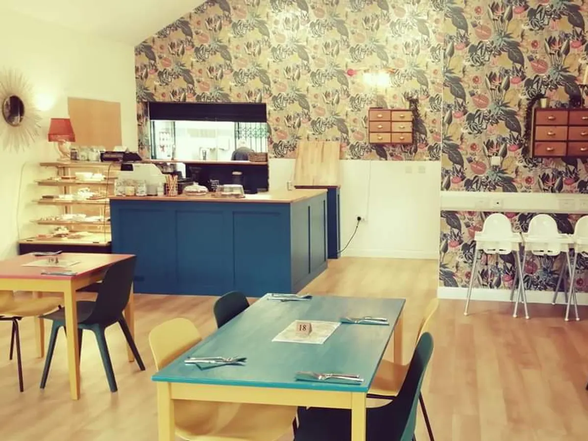 The interior of Cliffe Park Cafe showing a modern seating area with colourful patterned wallpaper, a blue counter with shelving, and wooden tables with chairs.