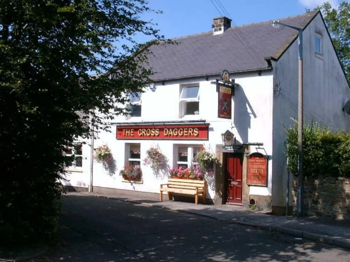 The front of the Cross Daggers pub on a clear day, showing the white-painted building with its red business sign, flowering planters, and red door.