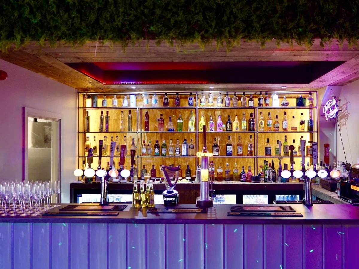 The bar area of Fox In The Box with extensive bottle display on backlit wooden shelves, purple mood lighting, and neon signage showcasing the nightlife offering.