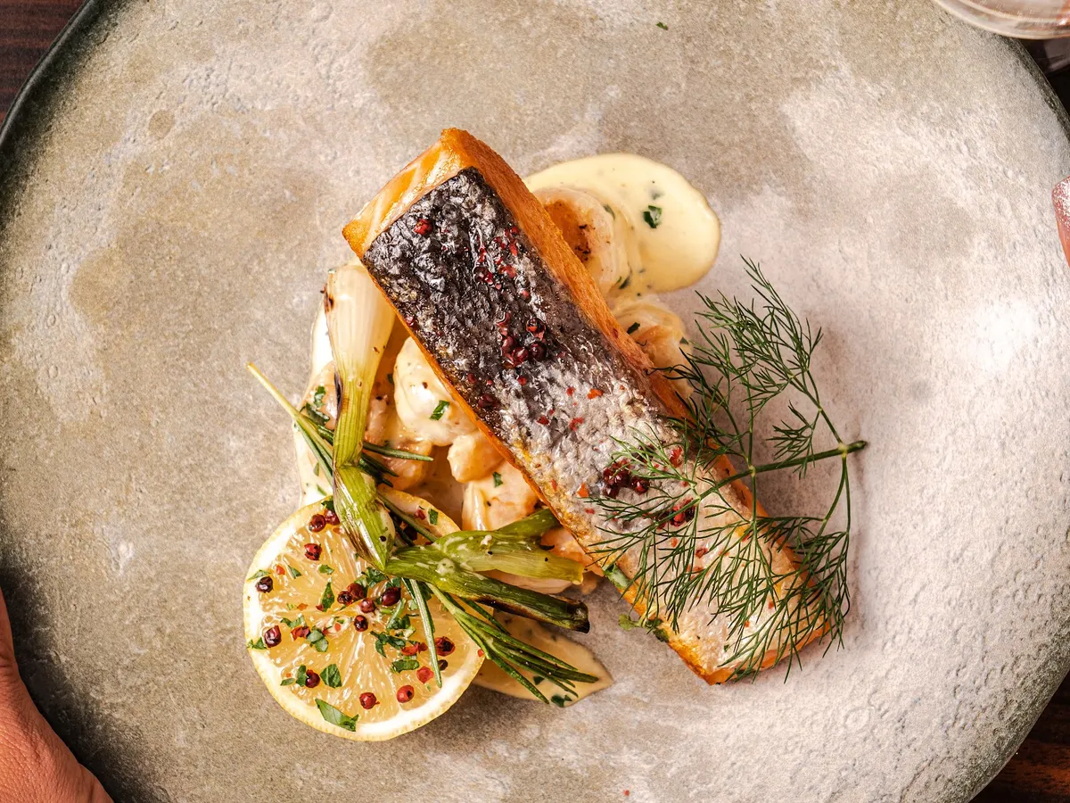 An elegantly plated main course with seared fish, herb garnish, lemon slice, and artistic sauce presentation on a pale ceramic dish.