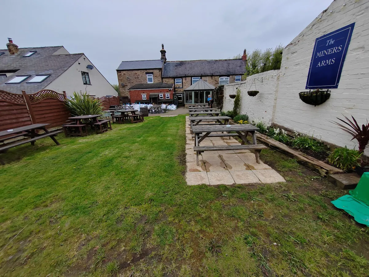 The outdoor patio and garden area of the Miners Arms with wooden picnic benches, green lawn, and pub sign visible on the building.