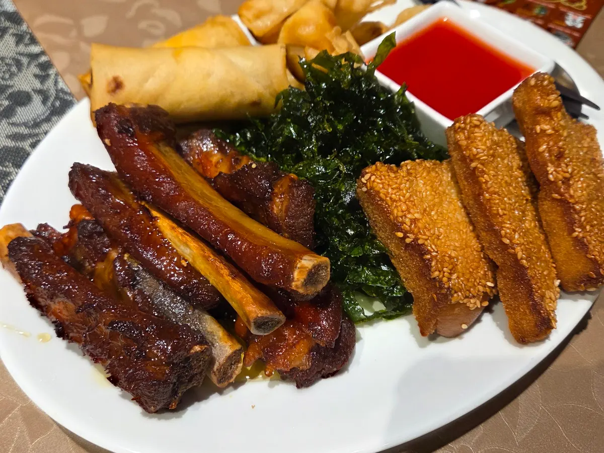 A white plate with roasted duck strips, spring rolls, sesame-coated items, and a small bowl of red sauce.