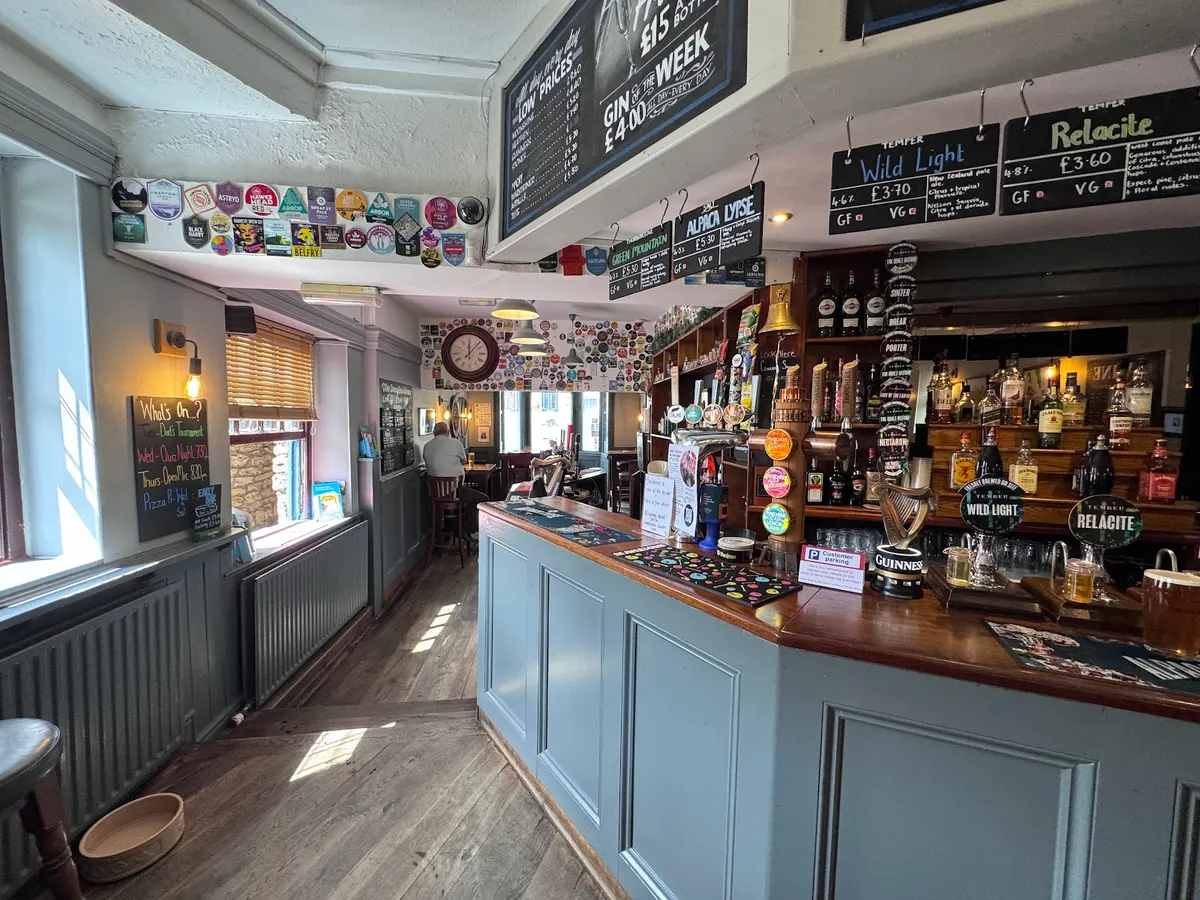 The interior of The Dronfield Arms bar, showing beer taps, branded pumps, bottles on shelves, and hanging brewery badges on the wall.