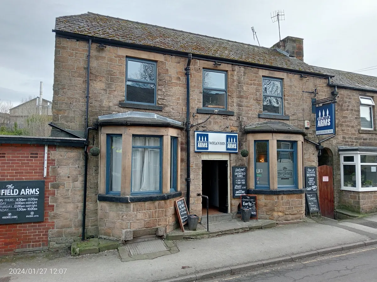 The stone building of The Dronfield Arms pub with blue and white signage clearly visible on the street, with benches outside.