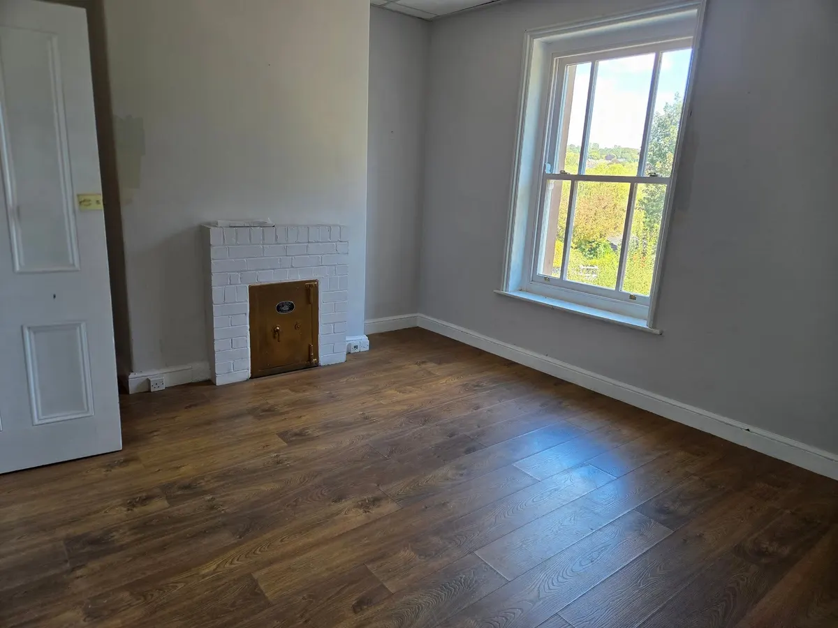 An empty room with a white-painted brick fireplace, a brass safe door and a tall sash window.