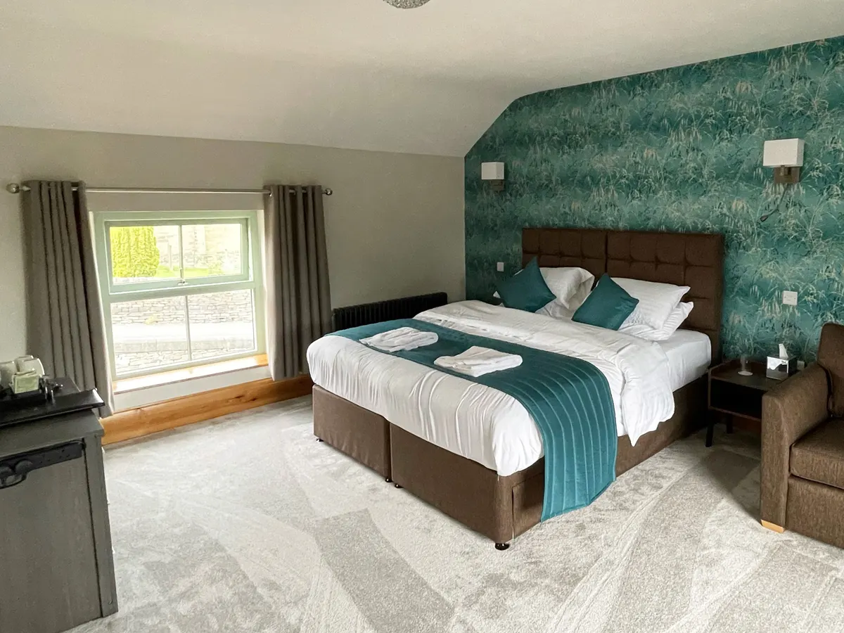 A hotel bedroom in The George And Dragon with a double bed, teal patterned wallpaper, and views through the window.