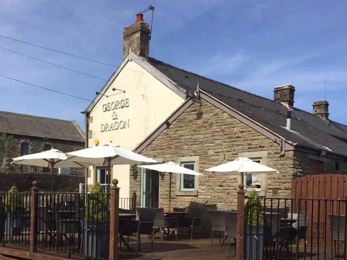 The stone building of The George And Dragon pub with the name clearly visible on a white gable wall, with outdoor seating area featuring umbrellas.