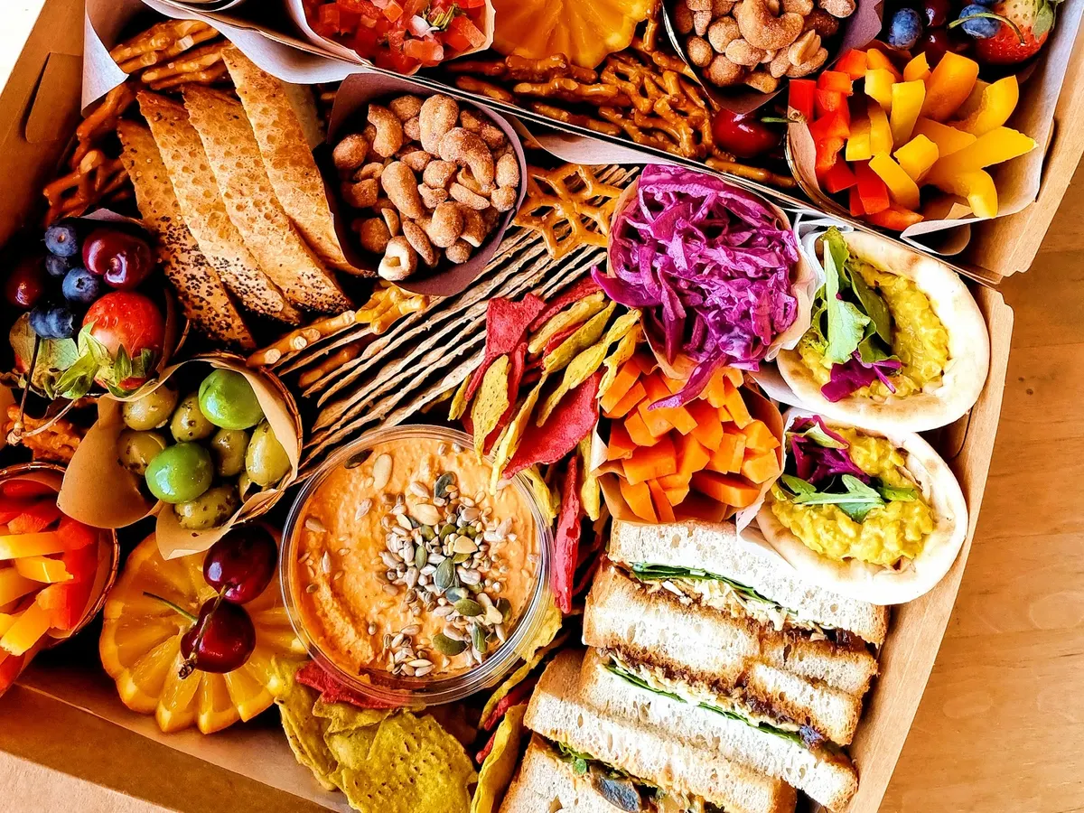 A wooden board displaying a spread of food from The Magic Sunflower including bread, spreads, vegetables, nuts, seeds, and hummus dips.