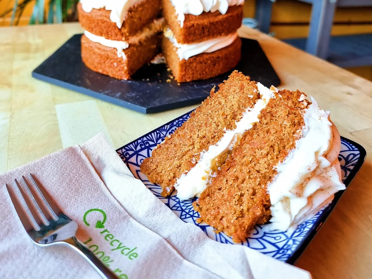 A slice of layered carrot cake with white frosting, served on a blue patterned plate at The Magic Sunflower.