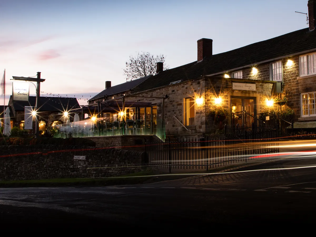 The stone front of the Peacock at Barlow lit up at dusk, with its terrace and hanging sign.