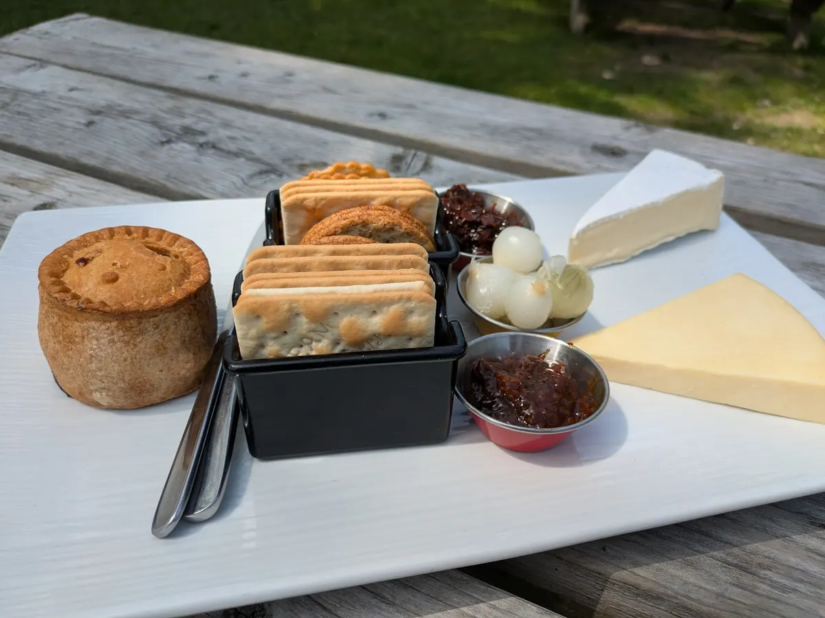 A charcuterie board at The Royal Oak featuring various cheeses, pies, jams, and relishes arranged on a white plate on a picnic table outdoors.