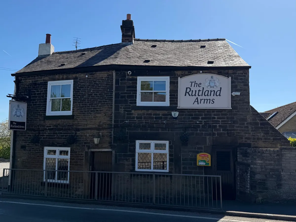 The front of The Rutland Arms pub showing the dark stone building, white-framed windows, and the Rutland Arms sign clearly visible.