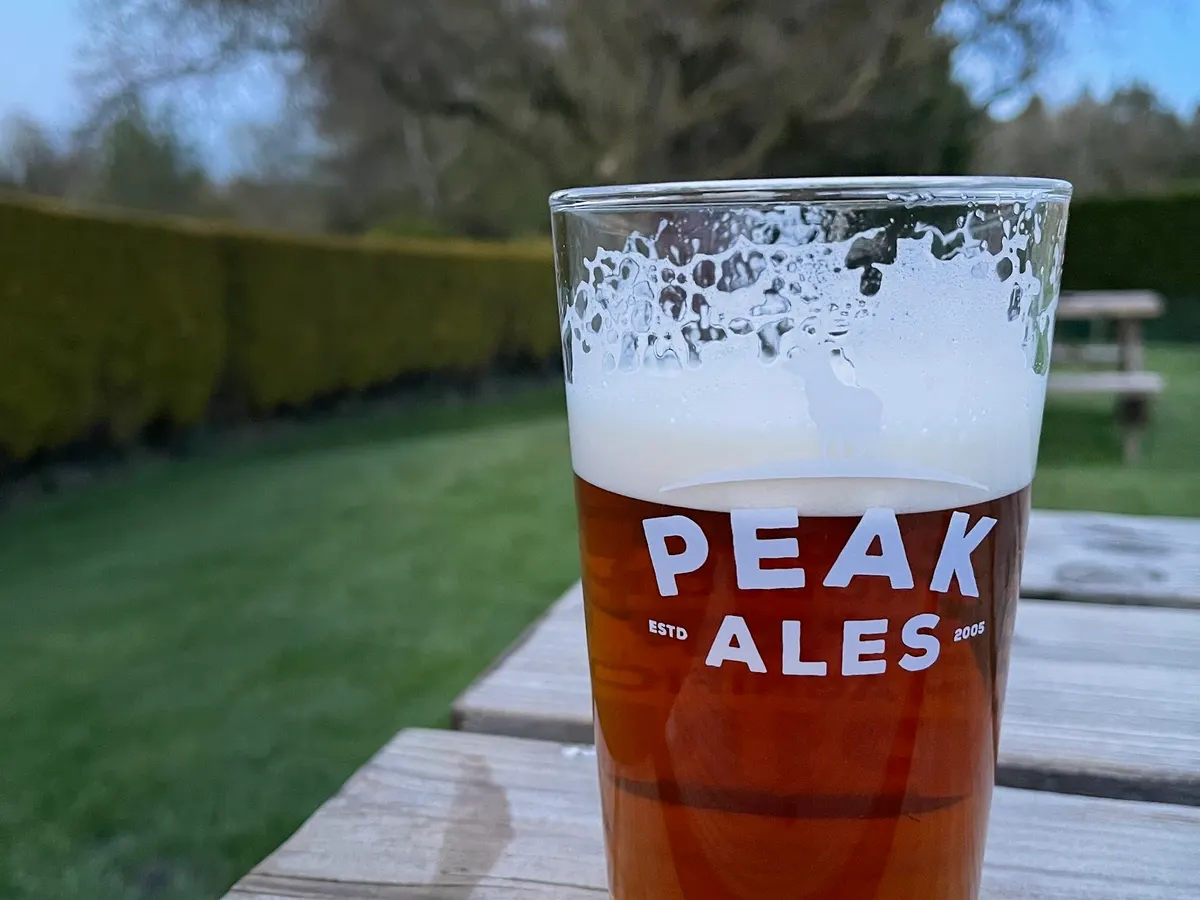 A glass of Peak Ales beer with the branded name visible, photographed on what appears to be an outdoor seating area with grass and hedgerow in the background.