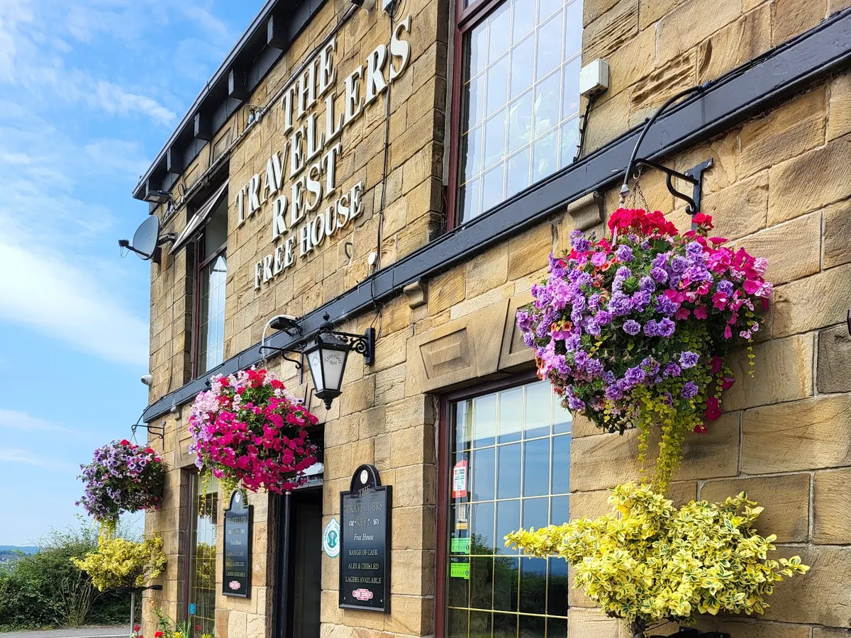 The Travellers Rest pub exterior with stone building, colorful hanging flower baskets, and the pub's sign visible.