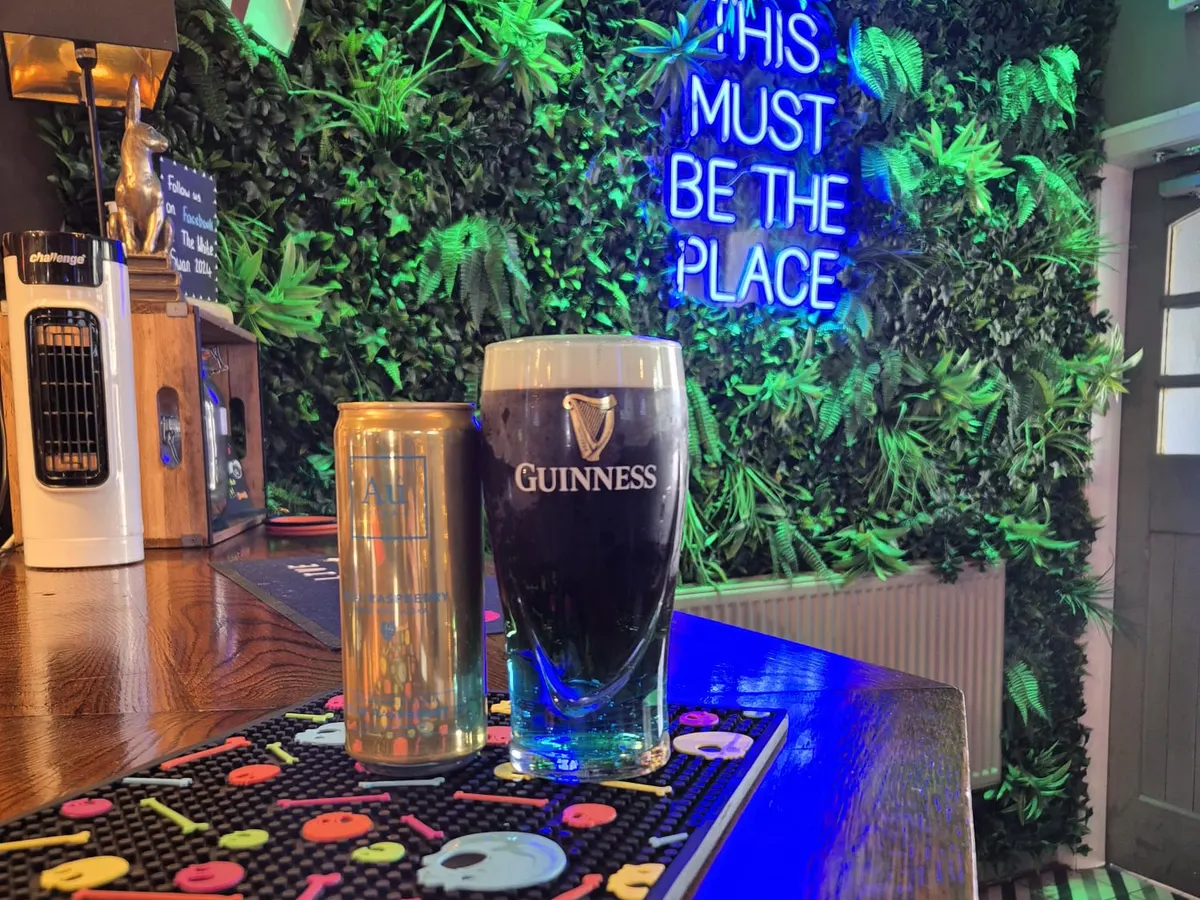 The White Swan interior bar area with a neon sign reading 'THIS MUST BE THE PLACE', Guinness glass, and green plant wall.