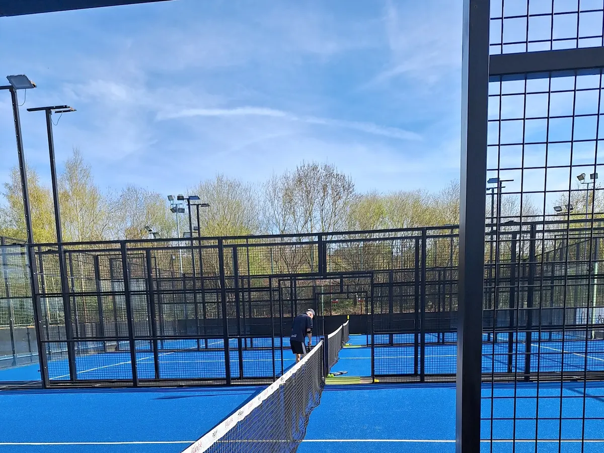 Blue padel courts under a blue sky, with black mesh fencing and a net in the foreground.
