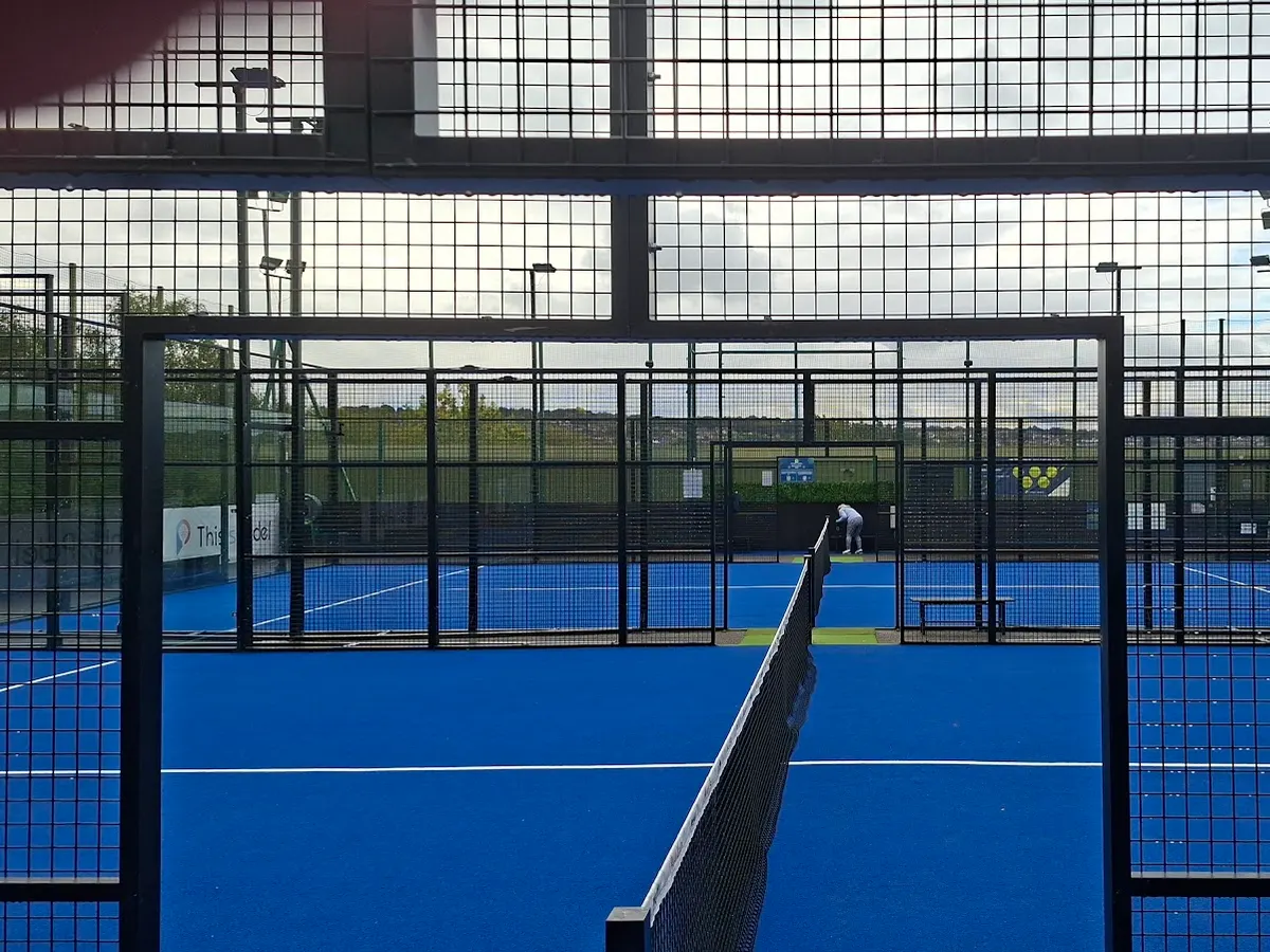 Blue padel courts seen through black mesh fencing, with the net in view.
