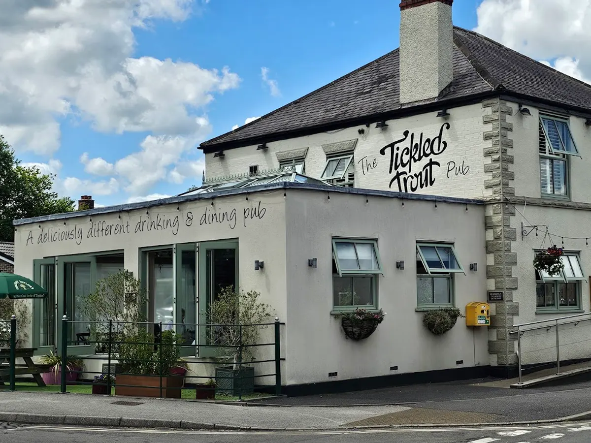 The Tickled Trout Pub exterior with white rendered and brick construction, green door, hanging flowers, and patio seating.