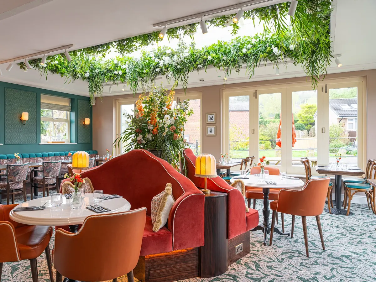 The Tickled Trout dining room with red velvet seating, hanging green plants, natural light from large windows, and set tables.
