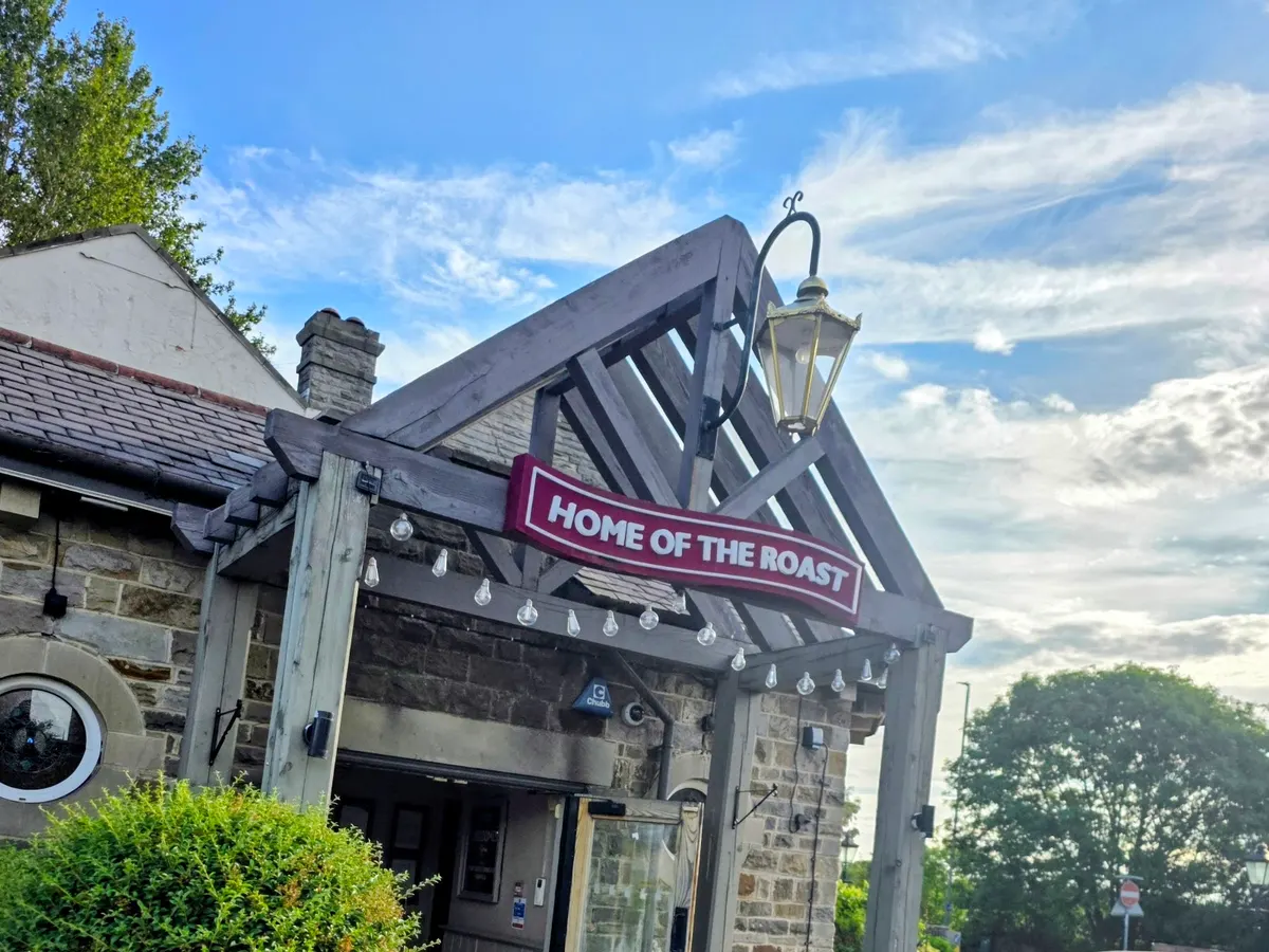 The front of Toby Carvery on a sunny day, showing the building with its distinctive wooden pergola and 'Home of the Roast' sign.